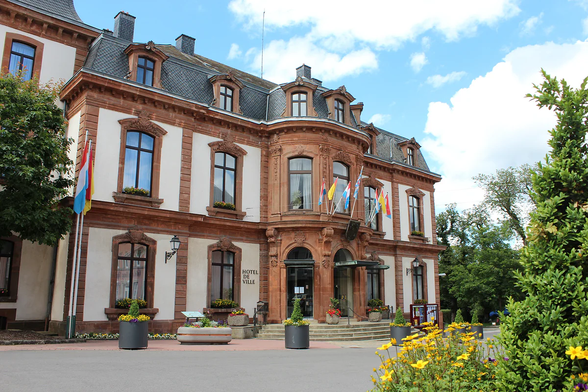 Wiltz town hall, a red and white stone building with a slate roof, flags and flowers at the entrance