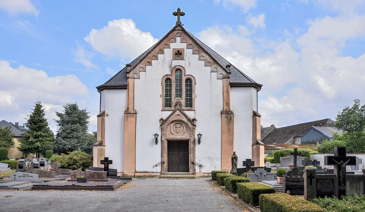 The white façade of Boulaide church with a reddish stone doorway, rose window and the cemetery crosses in front
