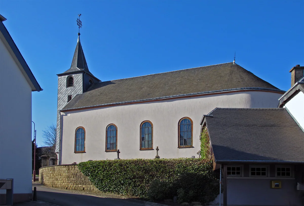 The church at Roodt, in the commune of Ell, white with a slate bell tower and arched windows, under a blue sky