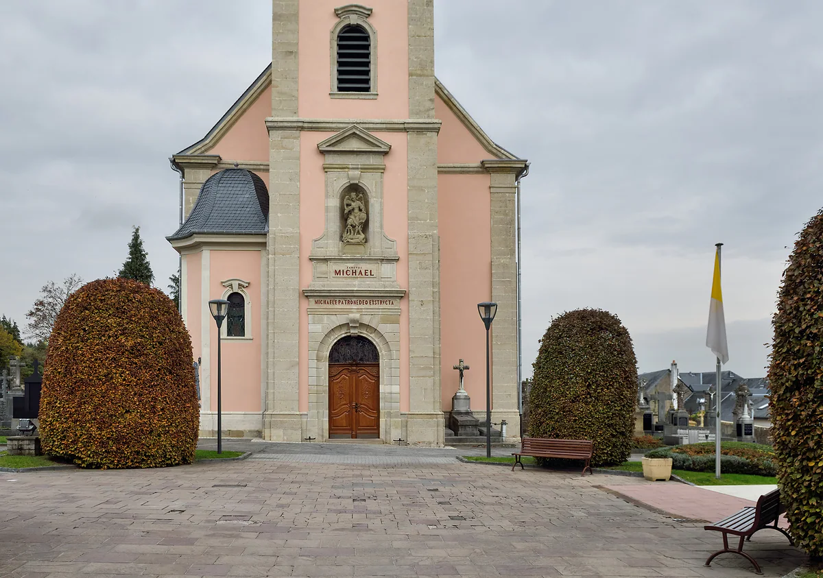 St Michael's Church in Mondorf-les-Bains, with a pink façade and tower, flanked by two rounded hedges