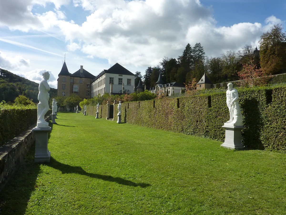 A grassy walk lined with white statues in the gardens at Ansembourg, with the new castle and hills behind