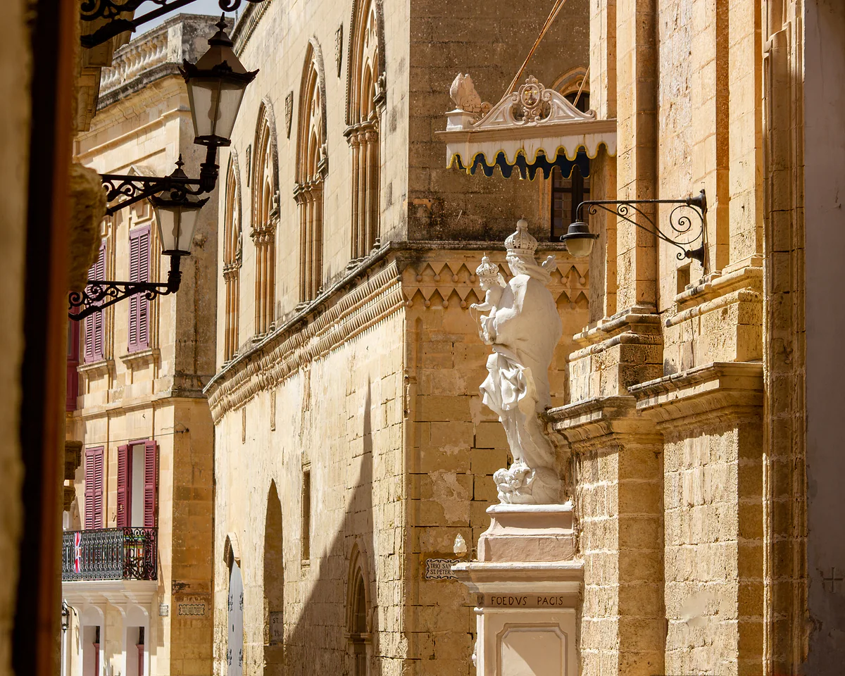 Una calle de Mdina con fachadas de piedra dorada, farolas de hierro, balcones y una estatua barroca en la esquina de un palacio.