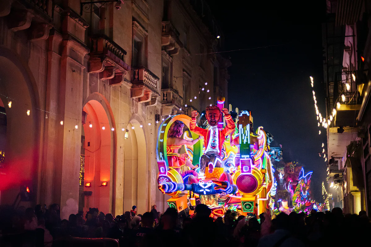 Carrozas del carnaval de Malta iluminadas de noche en una calle de La Valeta, con muñecos gigantes de colores y el público alrededor.