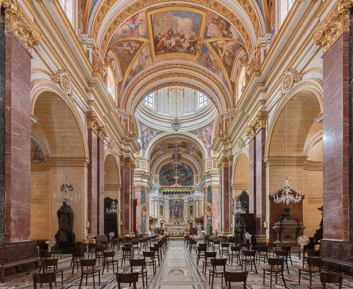 El interior de la catedral de San Pablo de Mdina, con pilastras de mármol rojo, bóveda pintada, lámparas de araña y el altar mayor al fondo.