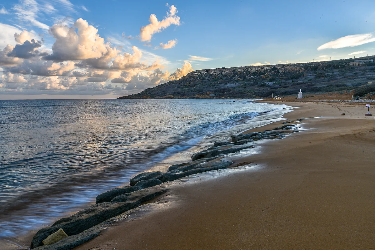 La playa de Ramla l-Ħamra, en Gozo, con su arena rojiza, las olas suaves en la orilla y la colina verde que cierra la bahía.