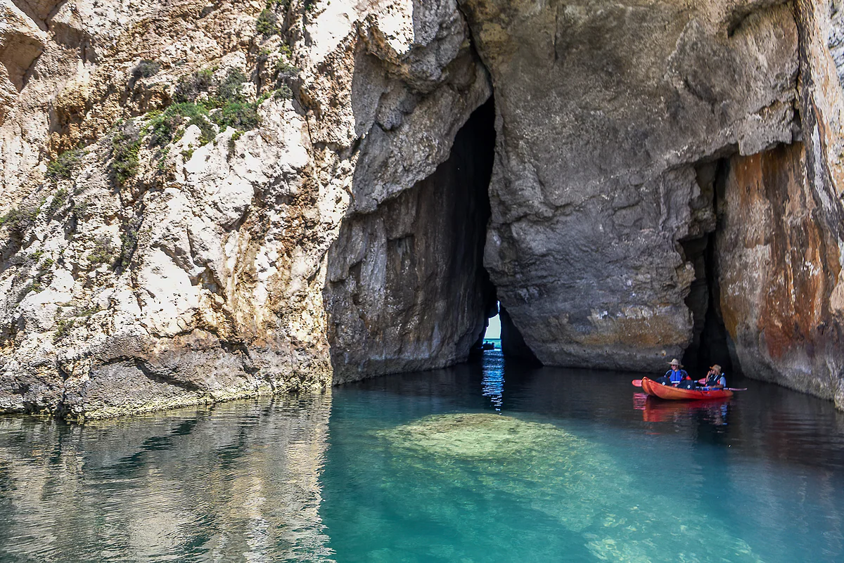 El túnel natural del Inland Sea de Dwejra, en Gozo, con el agua turquesa bajo la roca y un kayak entrando hacia el mar abierto.