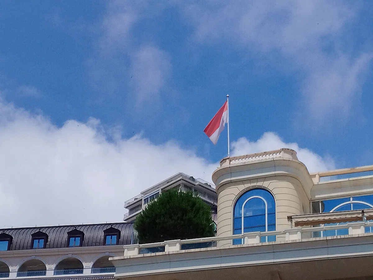 La bandera roja y blanca de Mónaco ondeando sobre un edificio del puerto bajo un cielo azul con nubes.