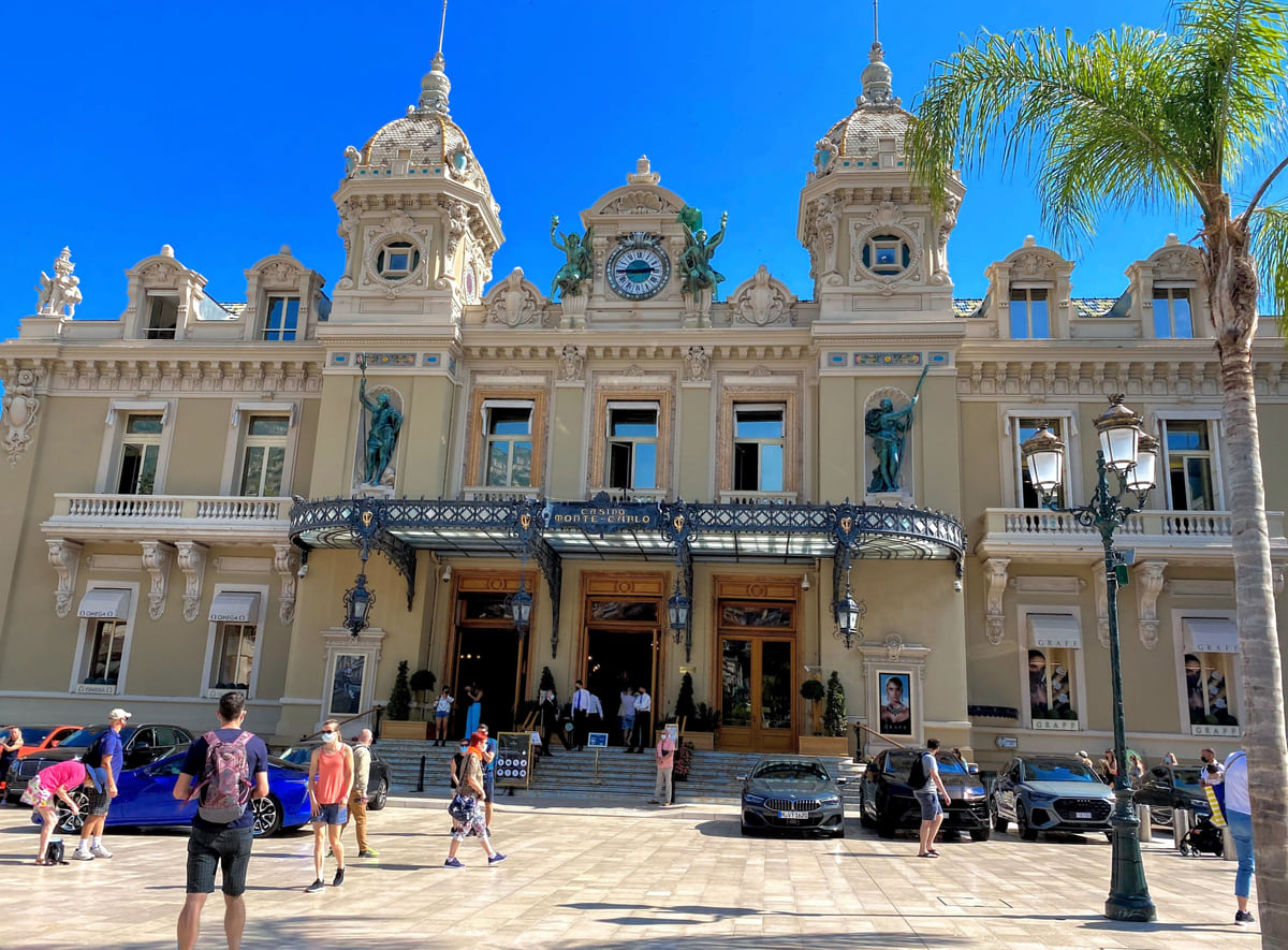 Facade of the Monte-Carlo Casino on Casino Square, with its two towers, the iron canopy and people strolling in front.