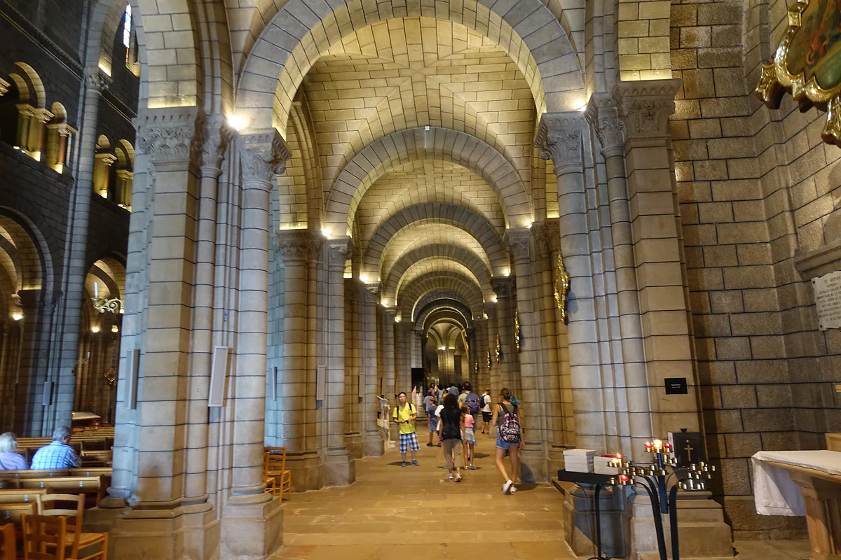 Deambulatorio de la catedral de Mónaco con sus arcos de piedra iluminados y visitantes recorriéndolo.