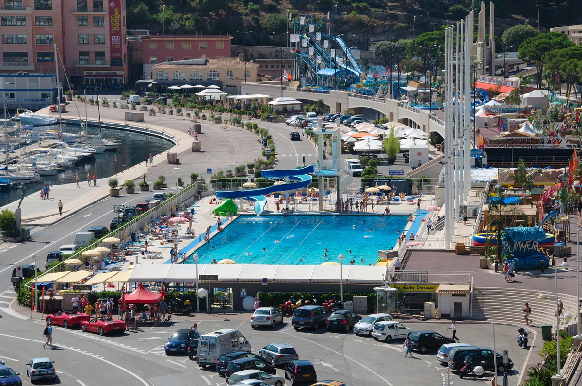 La piscina Rainiero III en el puerto de Hércules, llena de bañistas en verano, con su tobogán, el trampolín y los barcos amarrados a la izquierda.
