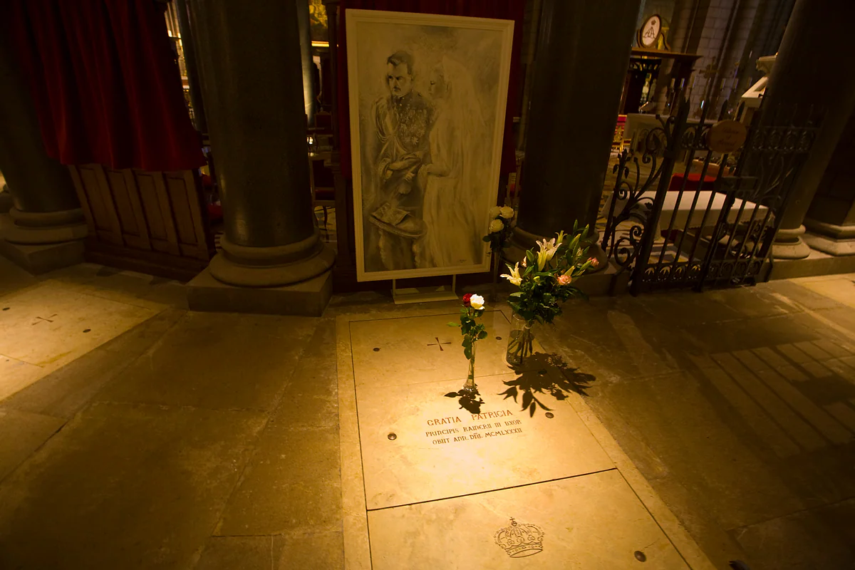 La lápida de la princesa Grace en el suelo de la catedral de Mónaco, con la inscripción «Gratia Patricia», flores y un retrato a lápiz detrás.