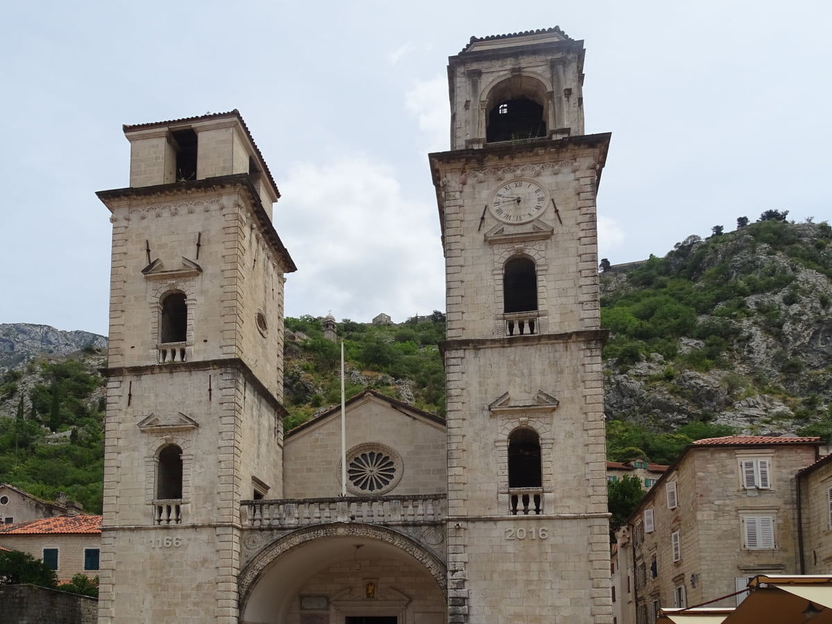 The front of St Tryphon's Cathedral in Kotor, with its two pale stone towers and rose window, below the mountains of the old town