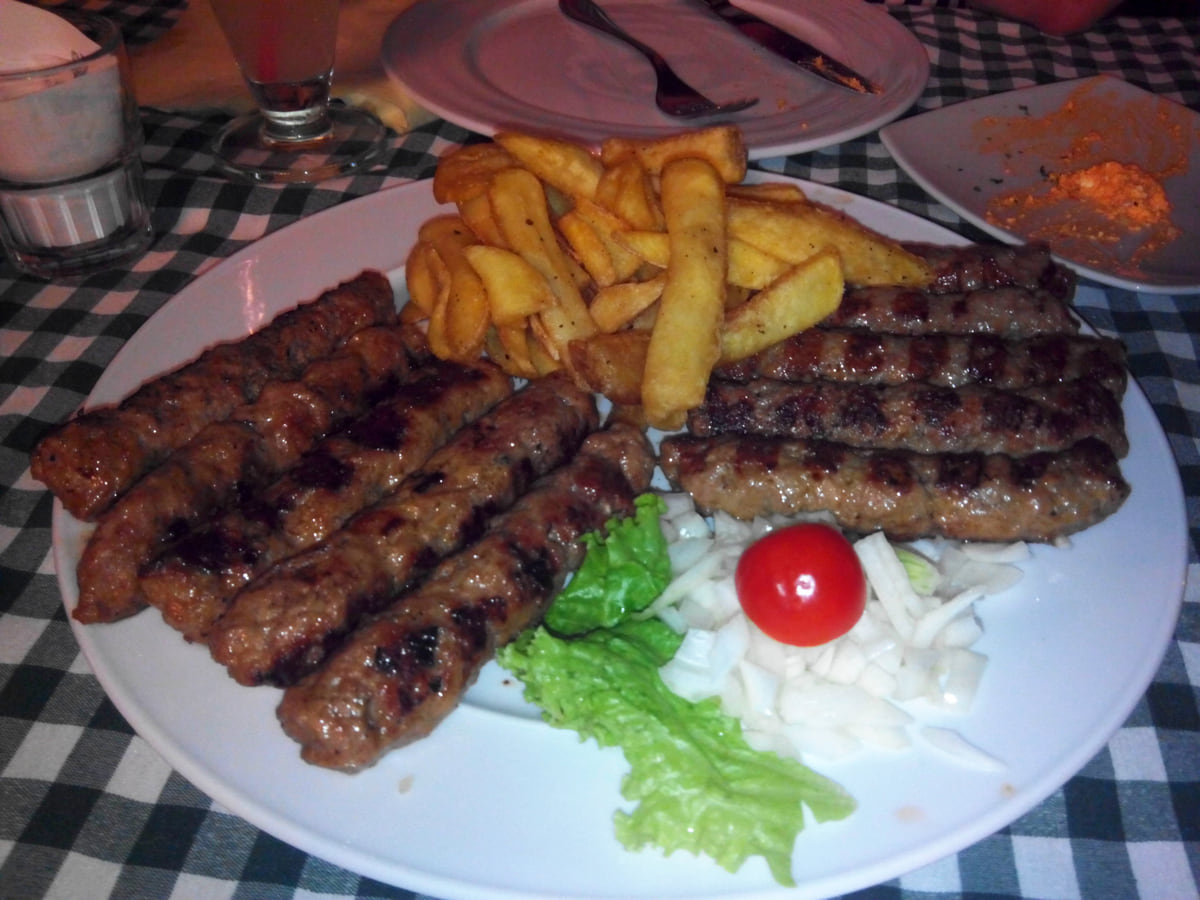 A plate of ćevapi with chips, chopped onion, lettuce and tomato on a checked tablecloth at a restaurant in Budva