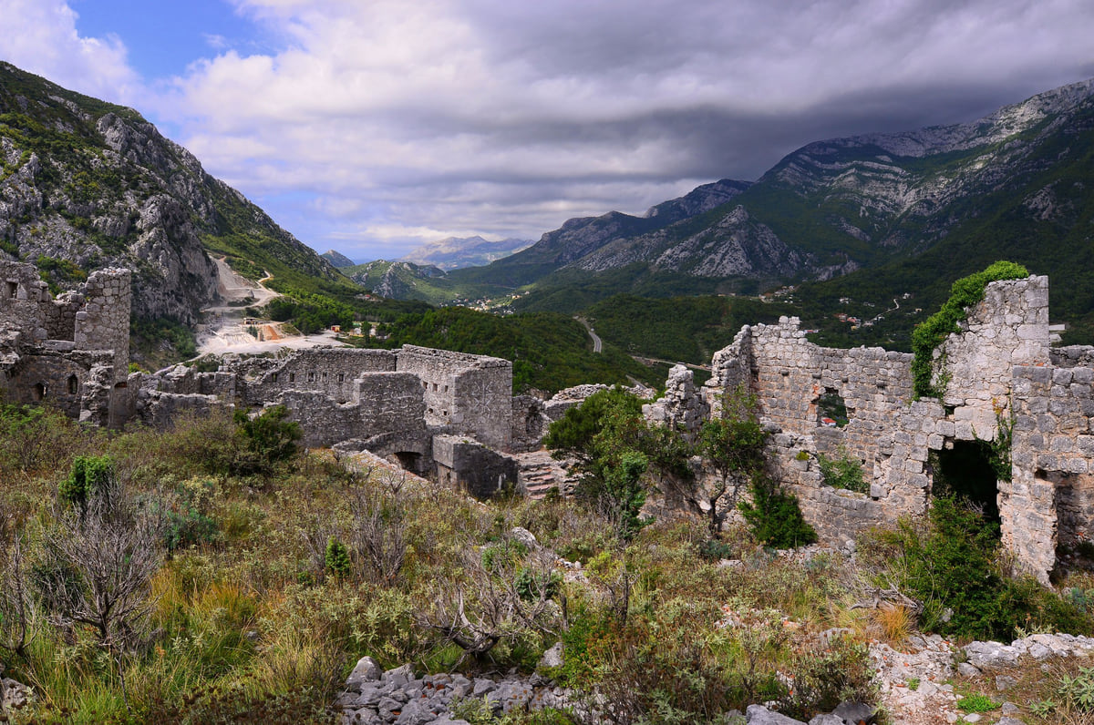 The ruined walls of Haj-Nehaj fortress among scrub, with the Sutomore valley and grey mountains under heavy cloud