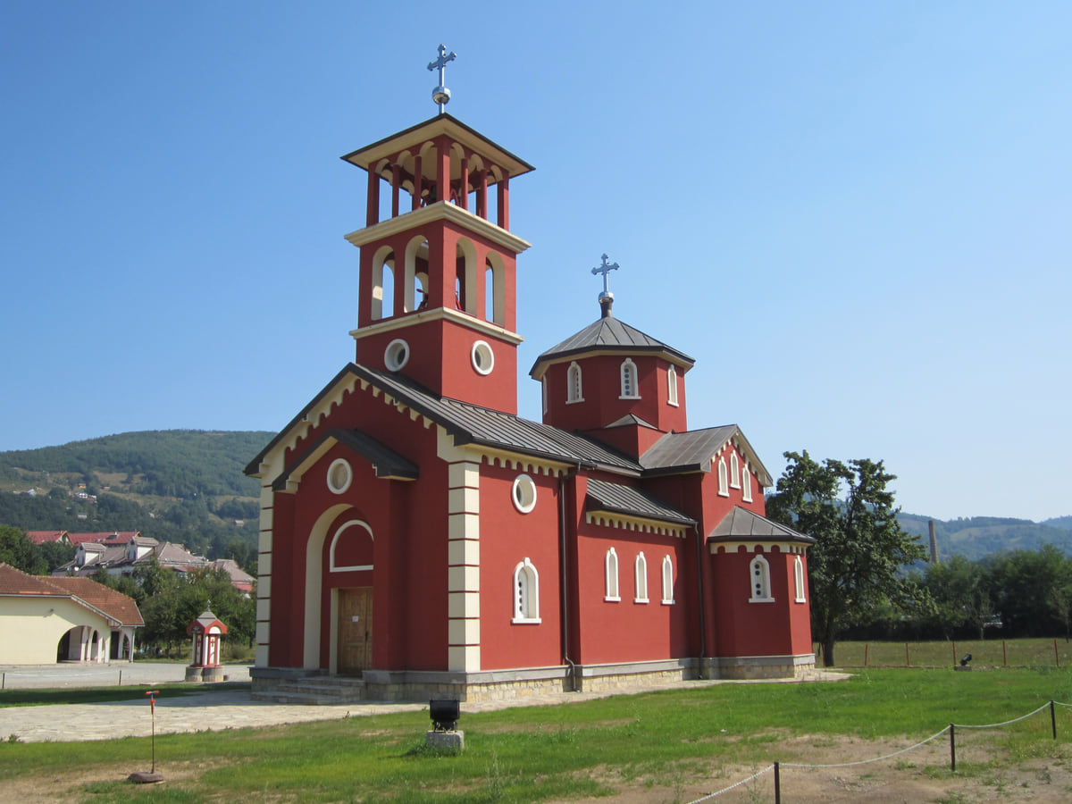 A red Orthodox church with a bell tower and dome in Mojkovac, on a green lawn with the wooded hills of the Tara valley behind