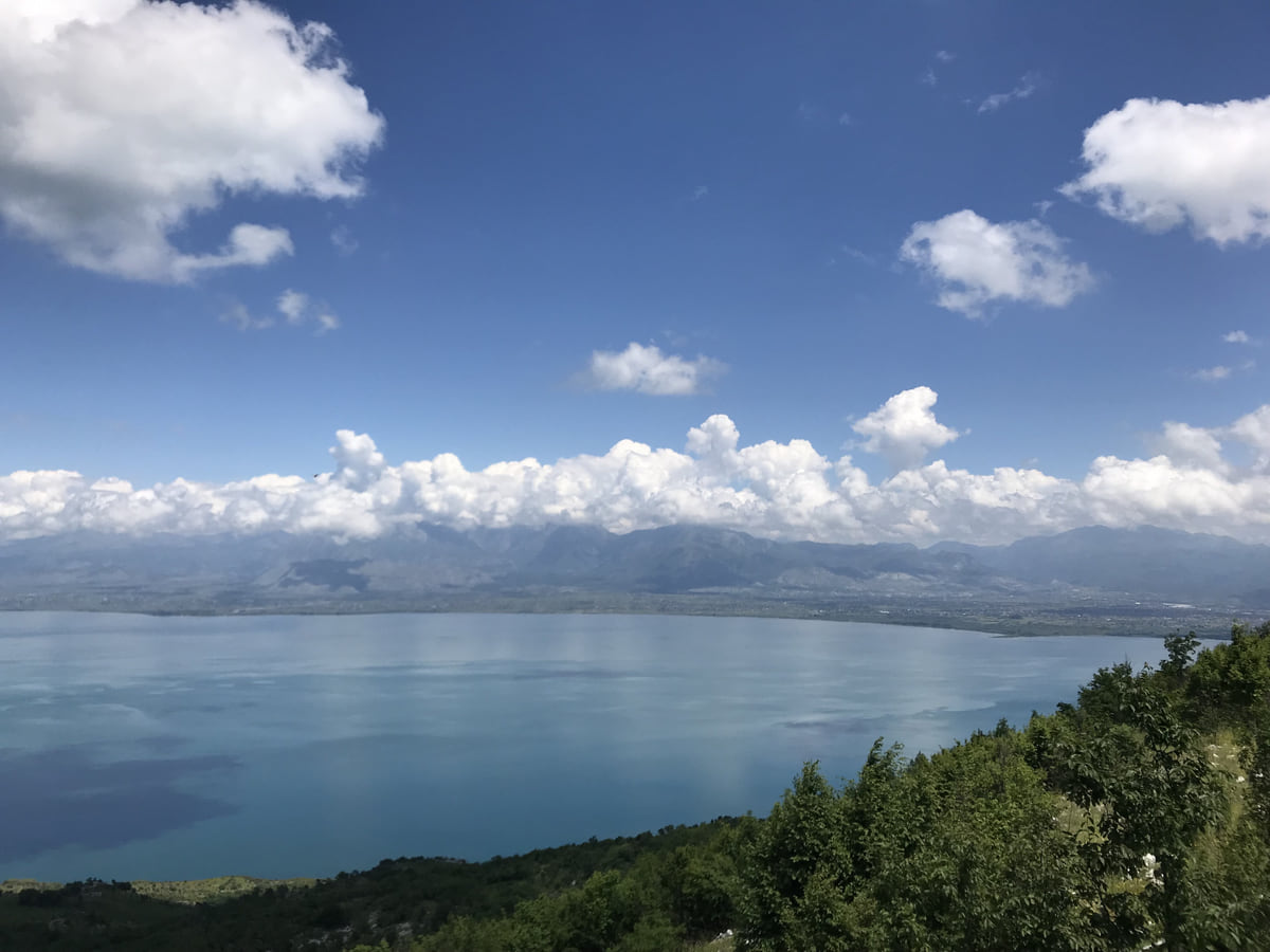 Lake Skadar from the Montenegrin shore, its blue water stretching to the mountains with white clouds on the horizon
