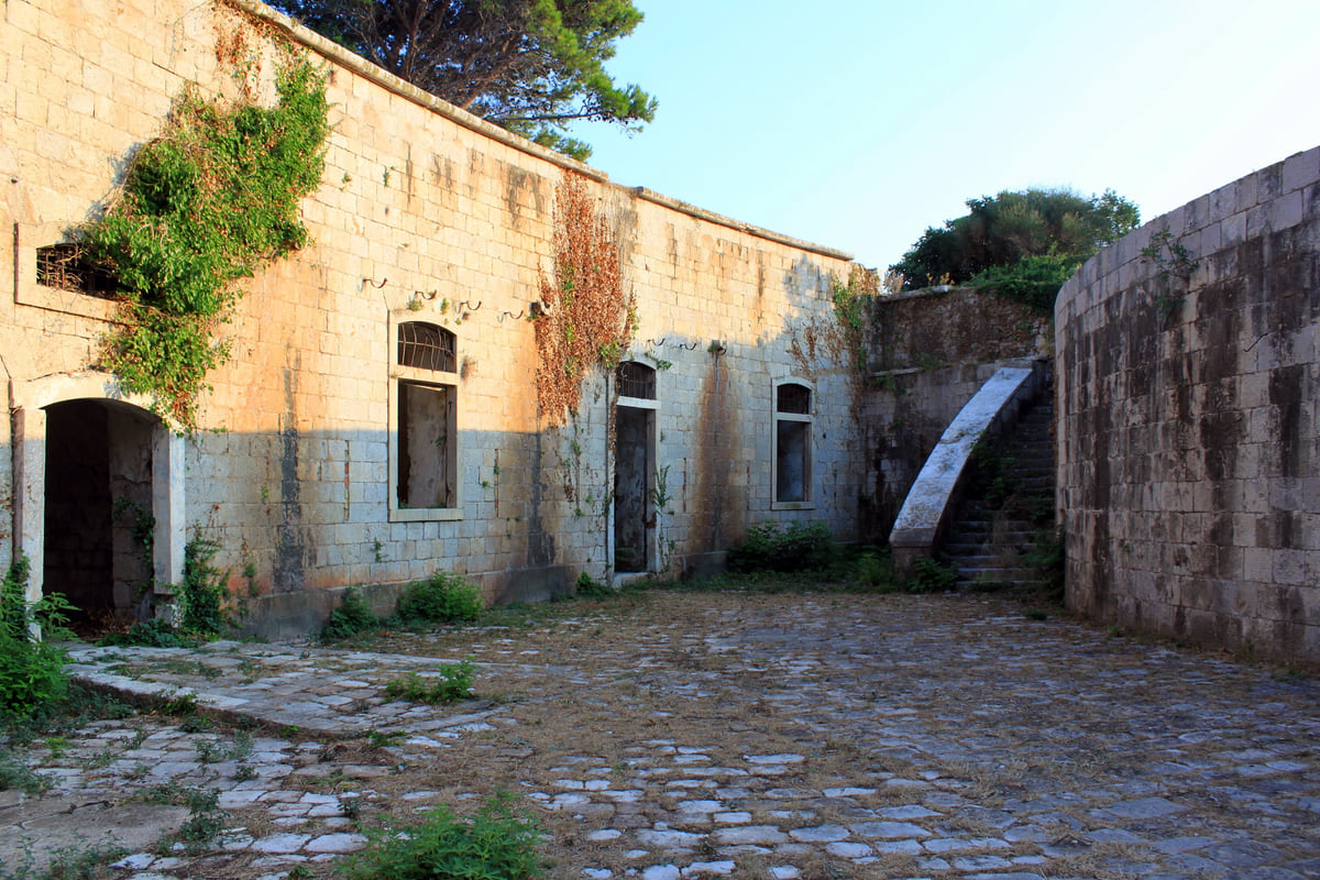 The cobbled courtyard of Mamula fortress, with stone walls, windows and plants growing on them, before its restoration