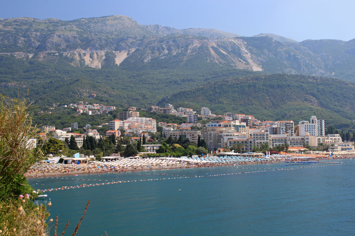Bečići beach with hotels lining the sand and mountains behind, seen from across the bay on a clear day