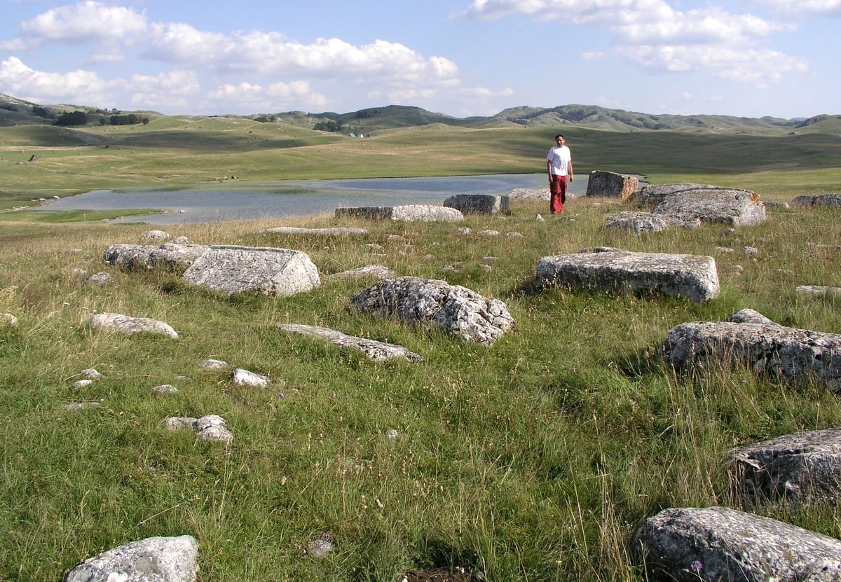 Medieval stećci tombstones scattered across a Durmitor meadow at Grčko groblje near Žabljak, with a lake behind
