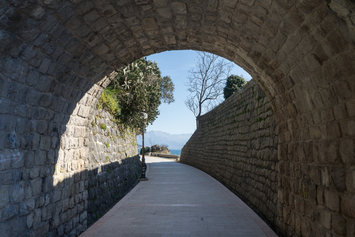 A stone tunnel on Herceg Novi's seafront promenade opening towards the sea, with a dressed-stone wall and trees beyond
