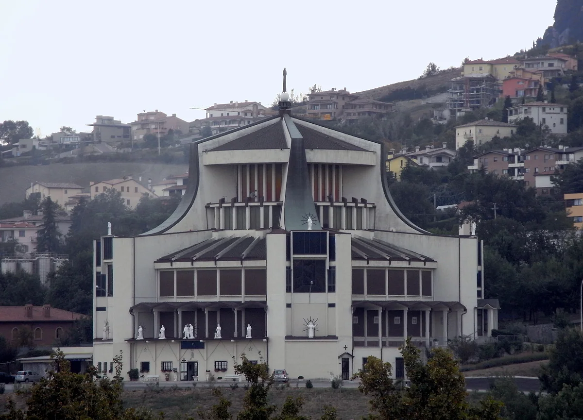 El santuario del Corazón Inmaculado de María de Valdragone, de fachada blanca y tejados en abanico, con las casas de la ladera detrás.