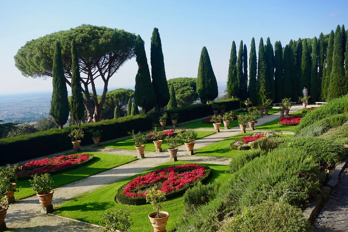 Parterres con flores rojas, macetas de limoneros, cipreses y pinos piñoneros en los jardines de Villa Barberini, con la llanura hacia el mar al fondo.