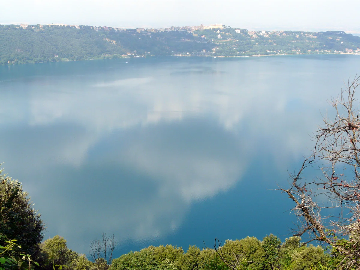 El lago Albano en calma reflejando las nubes, con el borde boscoso del cráter y el pueblo de Castel Gandolfo en lo alto de la orilla opuesta.